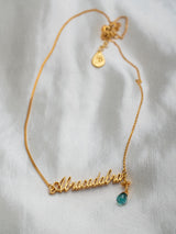 A gold-colored necklace handmade by Leeloobird Bali with the word 'ABRACADABRA' written in cursive and a small blue gemstone pendant attached to the end.